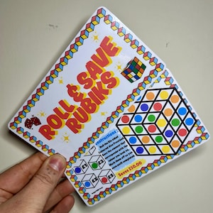 Op de afbeelding: Een kleurrijke spelkaart met instructies om een ​​zeszijdige dobbelsteen te gooien en het bedrag te sparen dat overeenkomt met de kleur waarop je landt. De kaart bevat een Rubik's Cube en een raster met gekleurde cirkels. De tekst "Roll & Save Rubiks" is gedrukt in grote, kleurrijke letters. De kaart vermeldt "Saves £50.00".