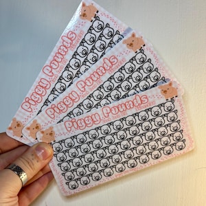 Puede incluir: Varias tarjetas rosas y blancas de "Piggy Pounds" se muestran en abanico. Cada tarjeta tiene una cuadrícula de caras de cerdos y el texto "Piggy Pounds" en rosa. Las tarjetas están diseñadas para rastrear el ahorro, cada tarjeta ahorra £50.