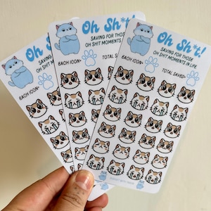 以下が含まれることがあります： 水色の猫のグラフィックと「Oh Sh*t! Saving for those Oh Shit Moments in Life」のテキストが入った3枚のステッカーシート。各シートには、さまざまな表情の猫の顔のアイコンが描かれています。ステッカーは光沢のある白い仕上がりです。