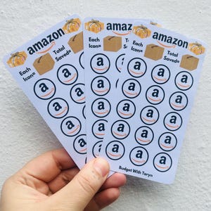 May include: Three white sticker sheets featuring the Amazon logo and text. Each sheet has the Amazon logo, a brown box graphic, and the text "Each Icon=" and "Total Saved=". The stickers are held in a hand.