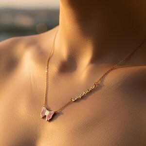 May include: A gold-colored necklace with a butterfly pendant and the name "Sophia" in cursive script. The butterfly has pink and white enamel detailing. The necklace is worn on a person's neck.