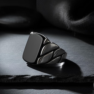 May include: A black signet ring with a rectangular face and a braided band design. The ring is made of a dark, possibly metal material, and sits on a dark surface. The ring's face reflects light, highlighting its polished finish.