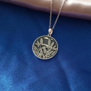 May include: A silver necklace featuring a circular pendant with an engraved wheat field design. The pendant has a detailed depiction of wheat stalks, grass, and clouds. The chain is a delicate silver link.
