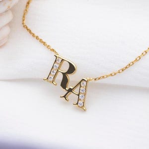 May include: A gold-colored necklace featuring the letters "R" and "A". Each letter is adorned with small, clear gemstones. The necklace is displayed on a white, textured surface, highlighting the jewelry's design and sparkle.