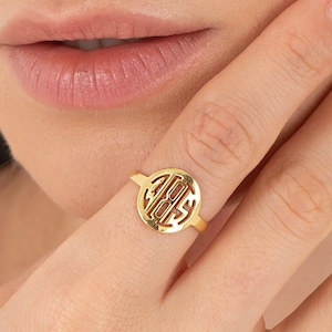 Puede incluir: Anillo de oro con un diseño circular que presenta un monograma estilizado. El anillo se lleva en un dedo.