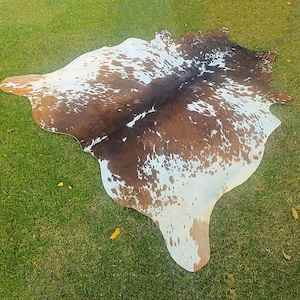以下が含まれることがあります： 茶色と白の斑点模様が自然なカウハイドラグ。ラグは不規則な形で、緑の草の上に置かれ、その有機的な形と質感を際立たせています。革の色のバリエーションは、素朴な美しさを生み出しています。