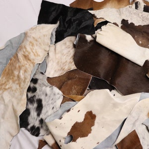 Puede incluir: Un montón de recortes de cuero de vaca de varios colores y patrones, incluyendo blanco, marrón, negro y moteado. Las pieles están dispuestas de forma aleatoria, con algunas superpuestas.