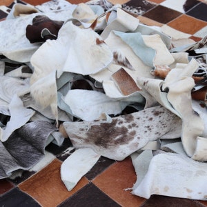 May include: A pile of white, brown, and black cowhide leather scraps. The leather is cut into various shapes and sizes. The leather is arranged on a checkered surface.