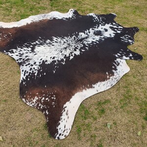 May include: A black and white cowhide rug with brown patches. The rug has a natural, irregular shape and is laid out on a green grassy surface.