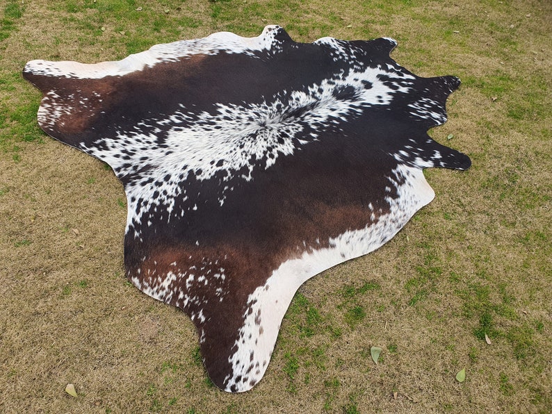 May include: A black and white cowhide rug with brown patches. The rug has a natural, irregular shape and is laid out on a green grassy surface.