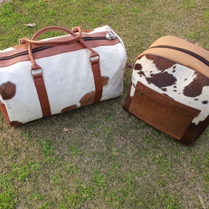 K&ouml;nnte beinhalten: Zwei braune und wei&szlig;e Rindledertaschen. Eine ist eine Reisetasche mit braunen Ledergriffen und -riemen. Die andere ist ein Rucksack mit einer braunen Lederklappe.