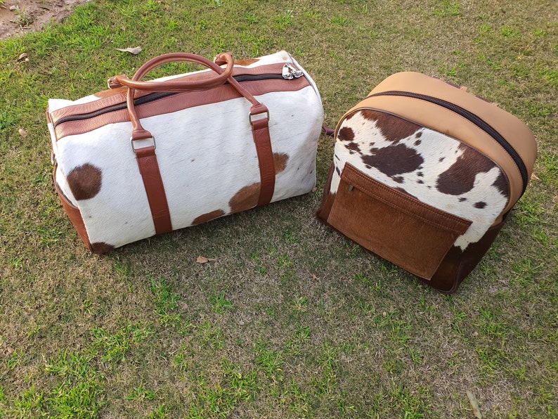 K&ouml;nnte beinhalten: Zwei braune und wei&szlig;e Rindledertaschen. Eine ist eine Reisetasche mit braunen Ledergriffen und -riemen. Die andere ist ein Rucksack mit einer braunen Lederklappe.