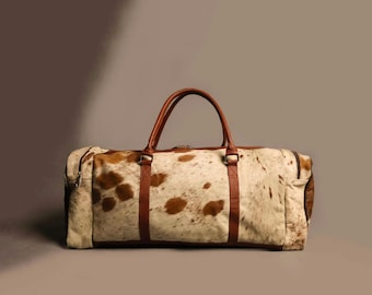 Könnte beinhalten: Eine große, rechteckige Reisetasche mit einem Kuhfellmuster in Braun- und Cremetönen. Die Tasche hat braune Ledergriffe und Akzente, einschließlich Riemen und einem Reißverschluss. Die Tasche ist für Reisen konzipiert.