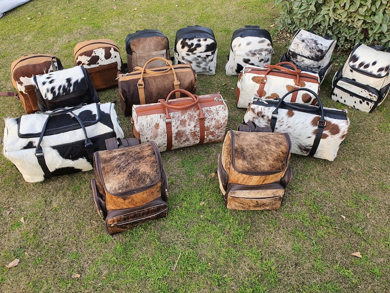 K&ouml;nnte beinhalten: Eine Sammlung von 14 Taschen aus Kuhfell in verschiedenen Stilen, darunter Rucks&auml;cke und Reisetaschen. Die Taschen sind alle braun und wei&szlig; mit einem gepunkteten Muster.
