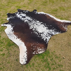 May include: A brown and white cowhide rug with a natural, spotted pattern. The rug is laid out on a green grassy surface.