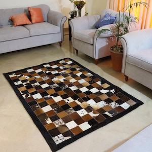 May include: A checkered patchwork rug made of cowhide in shades of brown, black, and white. The rug has a black border and is displayed on a light gray tiled floor in a living room setting.