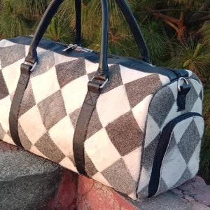 K&ouml;nnte beinhalten: Eine schwarze Leder-Reisetasche mit einem wei&szlig;en und grauen Rautenmuster. Die Tasche hat einen Rei&szlig;verschluss und eine kleine Tasche an der Seite.