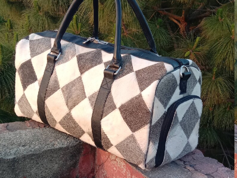 K&ouml;nnte beinhalten: Eine schwarze Leder-Reisetasche mit einem wei&szlig;en und grauen Rautenmuster. Die Tasche hat einen Rei&szlig;verschluss und eine kleine Tasche an der Seite.