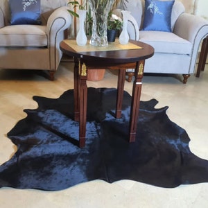 Pu&ograve; includere: Un tappeto in pelle di mucca nera con una tonalit&agrave; blu scuro, steso su un pavimento beige chiaro. Un tavolo rotondo in legno scuro con dettagli dorati si trova sopra il tappeto. Due vasi di vetro trasparente con fiori sono sul tavolo.