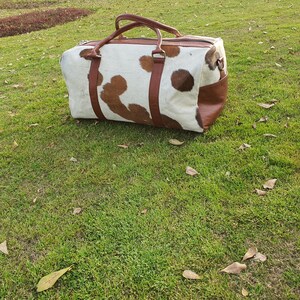 K&ouml;nnte beinhalten: Eine braune und wei&szlig;e Reisetasche aus Kuhfell mit braunen Lederelementen und Griffen.