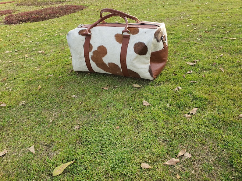 K&ouml;nnte beinhalten: Eine braune und wei&szlig;e Reisetasche aus Kuhfell mit braunen Lederelementen und Griffen.