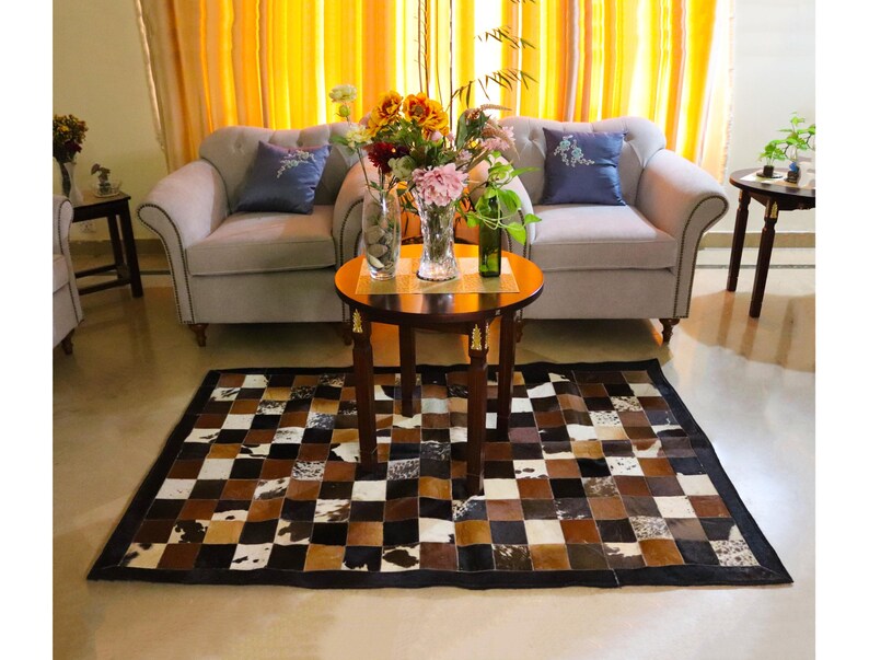 Op de afbeelding: Een bruin en wit patchwork koeienhuidkleed met een zwarte rand. Het kleed ligt in een woonkamer met twee witte banken, een salontafel en een vaas met bloemen.