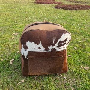 May include: A brown leather backpack with a white and brown cowhide pattern. The backpack has a large front pocket.