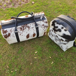 K&ouml;nnte beinhalten: Ein Set aus zwei Taschen aus Kuhfell. Die gr&ouml;&szlig;ere Tasche ist eine Reisetasche mit schwarzem Lederbesatz und einem Rei&szlig;verschluss. Die kleinere Tasche ist ein Rucksack mit schwarzem Lederbesatz. Beide Taschen haben ein wei&szlig;-braunes Kuhfellmuster.