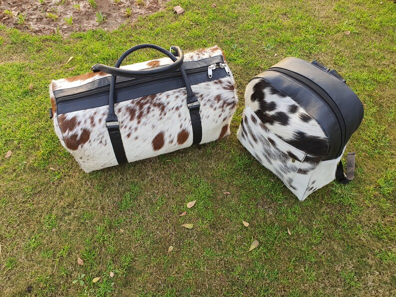 K&ouml;nnte beinhalten: Ein Set aus zwei Taschen aus Kuhfell. Die gr&ouml;&szlig;ere Tasche ist eine Reisetasche mit schwarzem Lederbesatz und einem Rei&szlig;verschluss. Die kleinere Tasche ist ein Rucksack mit schwarzem Lederbesatz. Beide Taschen haben ein wei&szlig;-braunes Kuhfellmuster.