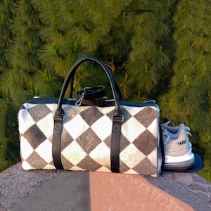 May include: A duffel bag with a black and white chequered pattern, black handles, and a black shoulder strap. A grey and white athletic shoe is placed in the side pocket of the bag. The bag is on a stone surface with a green background.