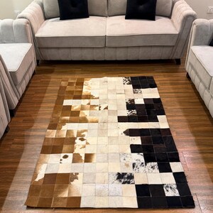 Peut inclure: Un tapis rectangulaire composé de carrés de peau de vache disposés en dégradé. Le tapis passe de motifs tachetés marron et blanc sur la gauche, à des tons crème et gris, pour finir par des carrés noirs et blancs unis sur le côté droit.