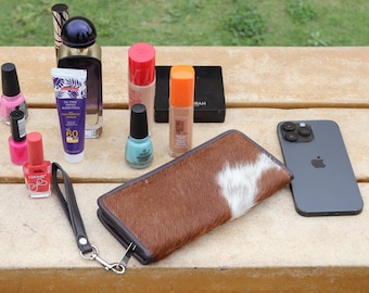 May include: A brown and white cowhide wallet with a black strap, a black iPhone with a camera bump, and various beauty products including nail polish, foundation, and sunscreen on a wooden surface.