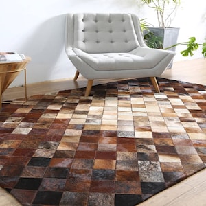 May include: A brown and beige patchwork cowhide rug with a diamond pattern. The rug is in a living room setting with a white chair and a wooden side table.