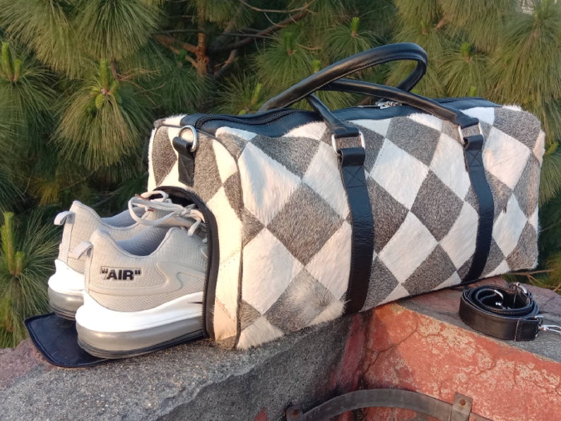 K&ouml;nnte beinhalten: Eine graue und wei&szlig;e karierte Ledertasche mit schwarzen Ledergriffen und einem schwarzen Ledertragegurt. Zwei graue Turnschuhe mit wei&szlig;en Sohlen und dem Wort "AIR" an der Seite befinden sich in der Tasche.