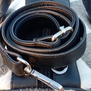 K&ouml;nnte beinhalten: Ein schwarzes Lederband mit silbernen Beschl&auml;gen ist auf einer wei&szlig;en und grauen Kuhhautfl&auml;che aufgerollt. Das Band ist f&uuml;r eine Tasche oder Handtasche gedacht.