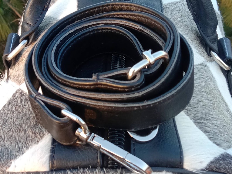 K&ouml;nnte beinhalten: Ein schwarzes Lederband mit silbernen Beschl&auml;gen ist auf einer wei&szlig;en und grauen Kuhhautfl&auml;che aufgerollt. Das Band ist f&uuml;r eine Tasche oder Handtasche gedacht.