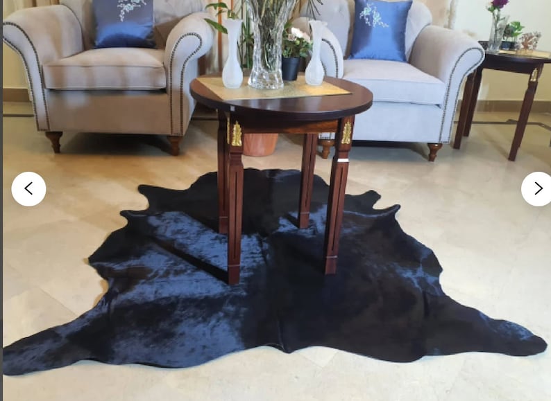 Pu&ograve; includere: Un tappeto in pelle di mucca nera con una tonalit&agrave; blu scuro, steso su un pavimento beige chiaro. Un tavolo rotondo in legno scuro con dettagli dorati si trova sopra il tappeto. Due vasi di vetro trasparente con fiori sono sul tavolo.