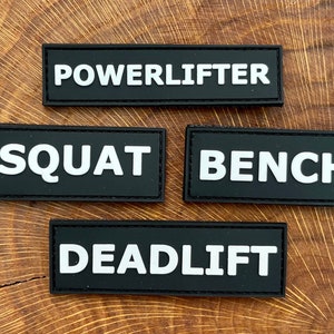 May include: Four black rectangular patches with white text. The patches read "POWERLIFTER", "SQUAT", "BENCH", and "DEADLIFT".
