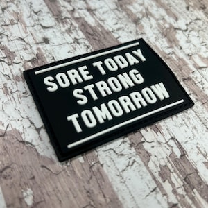 May include: Black and white motivational patch with the text "Sore Today Strong Tomorrow".