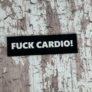 Könnte beinhalten: Ein schwarzer rechteckiger Aufnäher mit den Worten "FUCK CARDIO!" in fetten weißen Buchstaben. Der Aufnäher befindet sich vor einem strukturierten, verwitterten und abgenutzten Hintergrund mit Braun- und Weißtönen.