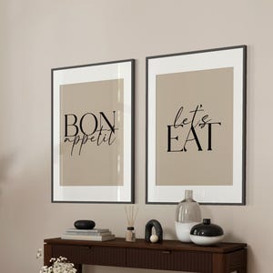 Peut inclure: Deux impressions encadrées avec des cadres noirs et des fonds beiges. L'impression de gauche dit "Bon appétit" en texte noir. L'impression de droite dit "Let's Eat" en texte noir.