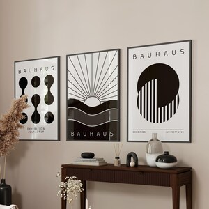 Puede incluir: Tres impresiones de arte minimalistas en blanco y negro con el texto "Bauhaus" y diseños geométricos. Las impresiones están colgadas en una pared beige claro sobre una consola de madera marrón oscuro con una mesa de café de vidrio en frente.