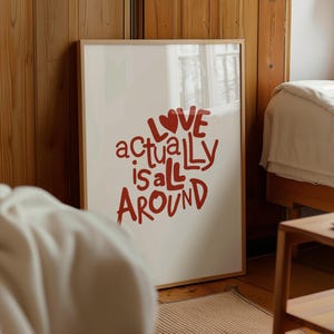 May include: A white framed poster with the text "Love actually is all around" in red lettering with a red heart symbol. The poster is hanging on a wooden wall.