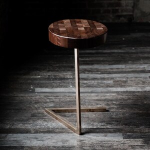Puede incluir: Una mesa auxiliar moderna con una tapa redonda de madera con diseño de mosaico. La mesa tiene una sola pata de metal delgada, de color dorado, que se conecta a una base triangular. La mesa está sobre un fondo oscuro.