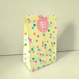 Könnte beinhalten: Eine gelbe Papiertüte mit bunten Tupfen. Ein rosa runder Aufkleber mit den Worten "happy happy happy" ist mit einer Schnur befestigt. Die Tüte ist oben gefaltet und gebunden.