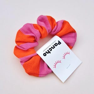 Könnte beinhalten: Ein farbenfrohes Haargummi mit abwechselnden rosa und orangefarbenen Streifen. Ein weißes Etikett mit dem Wort "pancho" in Schwarz, rosa Wimpern und den Worten "LOVELY HANDMADE" ist angebracht.