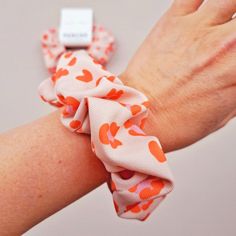 K&ouml;nnte beinhalten: Ein rosa und orangefarbenes Haargummi mit Leopardenmuster. Das Haargummi ist um ein Handgelenk gebunden.