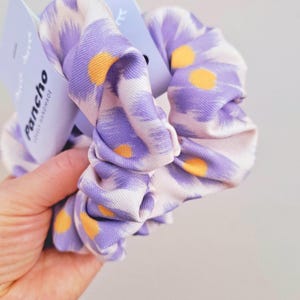 Könnte beinhalten: Zwei lila und weiße Haargummis mit gelbem Blumenmuster. Die Haargummis werden von der Firma "Pancho" hergestellt und als "lokal handgefertigt" beschrieben.