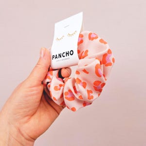 K&ouml;nnte beinhalten: Ein pinkfarbener und orangefarbener Scrunchie mit Leopardenmuster und einem wei&szlig;en Etikett mit der Aufschrift "PANCHO LOVELY HANDMADE" und einem gezeichneten Wimpernpaar.