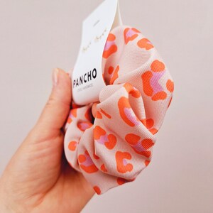 K&ouml;nnte beinhalten: Ein pink- und orangefarbener Scrunchie mit Leopardenmuster und einem wei&szlig;en Etikett mit der Aufschrift "PANCHO" und "SPICY HANDMADE".